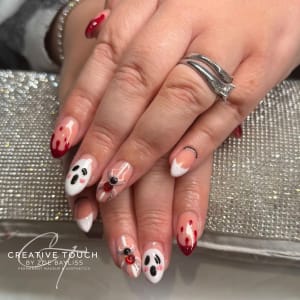 Close-up of hands with almond-shaped nails featuring various cute and spooky Halloween designs, including ghosts with rosy cheeks, red blood drips on white tips, a spider on a web, and a skeletal finger bone design on a nude base.