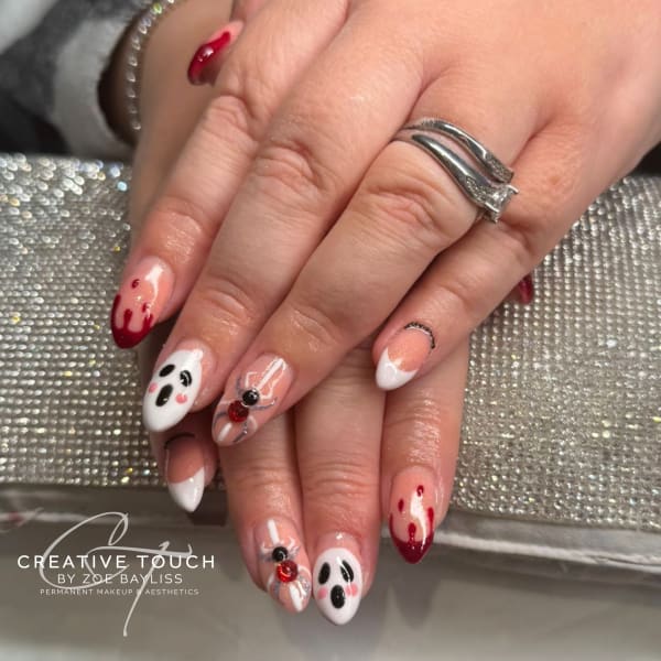 Close-up of hands with almond-shaped nails featuring various cute and spooky Halloween designs, including ghosts with rosy cheeks, red blood drips on white tips, a spider on a web, and a skeletal finger bone design on a nude base.