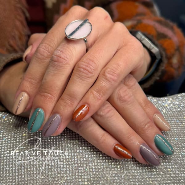 Close-up of hands with short, oval nails featuring a mixed color manicure in warm, earthy autumn shades: burnt orange, dusty mauve/purple, deep teal/green, and beige, with a rose gold glitter stripe accent on all coloured nails.