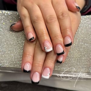 Close-up of hands with oval-shaped nails featuring elegant black French tips on a nude base. The ring finger stands out with a white French tip accented by a small, adorable black and white ghost motif.
