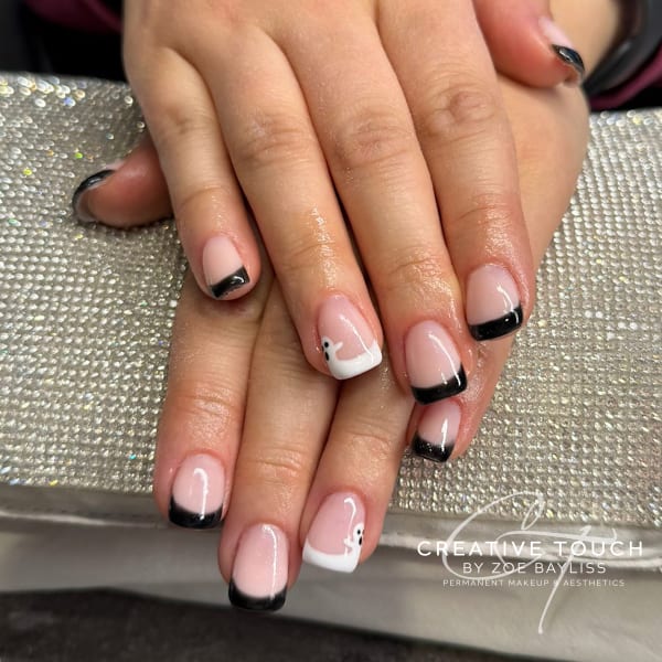 Close-up of hands with oval-shaped nails featuring elegant black French tips on a nude base. The ring finger stands out with a white French tip accented by a small, adorable black and white ghost motif.
