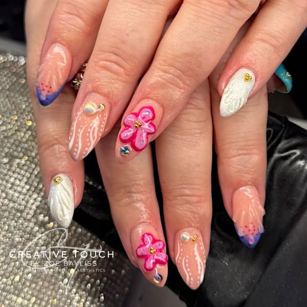 Close-up of hands with long, almond-shaped nails featuring a highly detailed, whimsical design on a nude-pink base. Elements include: 3D sculpted pink flowers with rhinestones, fine white lace/line work resembling a veil, curved French tips in deep purple/blue with red/pink dots, and shiny white textured accent nails resembling feathers or shells with gold accents.