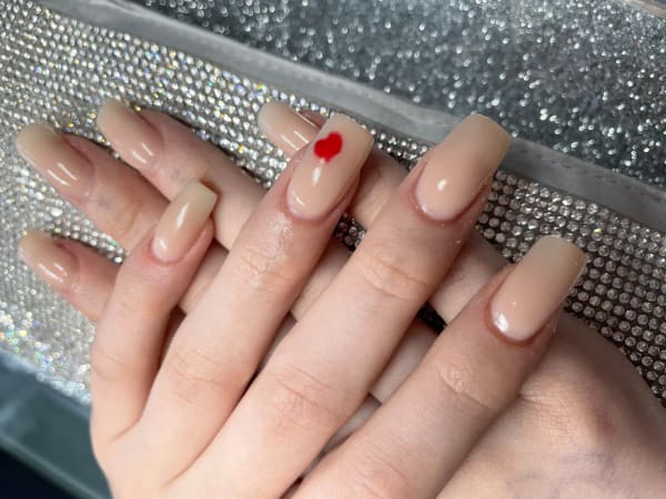 Close-up of a set of acrylic nails by Grace. The nails are beige/cream and boast a little red love heart on one nail. The nails are fairly long.
