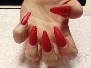 Close-up of a set of Stiletto-shaped acrylic nails. These are red with glitter bits and quite long. The shape is very pointy, like a stiletto. The hand is tilted up towards the camera, palm up. The background is a manicure towel.