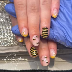 Close-up of a client's nails featuring glittery yellow tips with hand-painted bumble bees buzzing amongst delicate wildflowers.