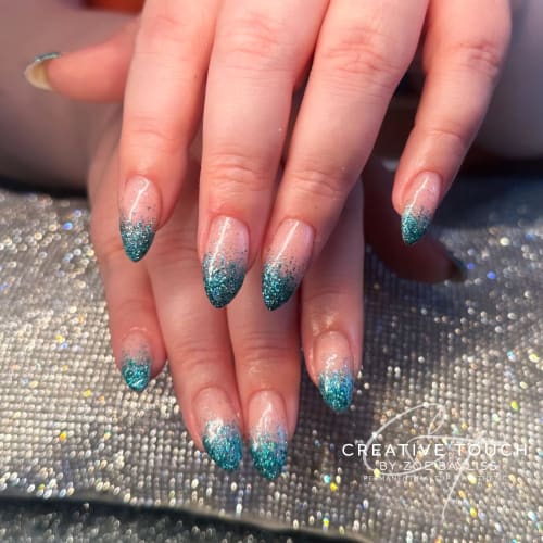 A close-up of hands with almond-shaped nails featuring a gradient from deep blue glitter to a nude basecoat.