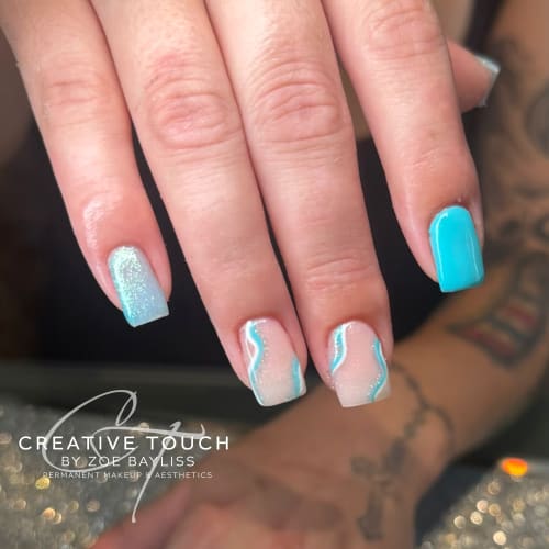 A close-up of hands with square nails featuring a mix of blue glitter, solid blue, and a blue and white wave design on a nude basecoat, reminiscent of a blue sky with trailing clouds.