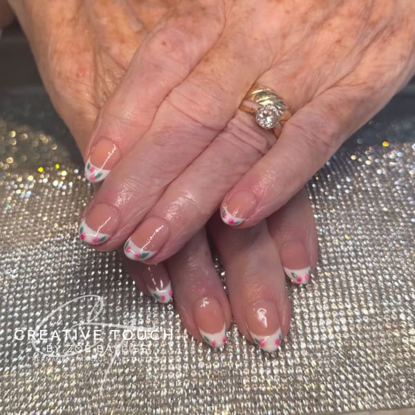 A close-up of hands with oval nails featuring a classic French tip design with two cherries and a green leaf on each tip.