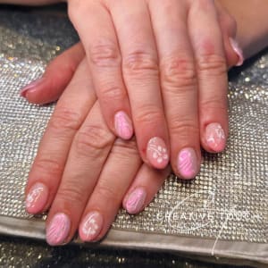A close-up of hands with oval nails featuring a pink swirl design on a nude basecoat with white floral accents.