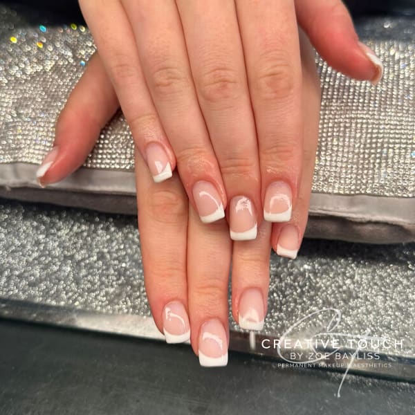 A close-up of hands with square nails featuring a classic French tip design with a white tip and a nude basecoat.