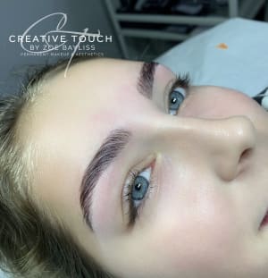 Portrait of our client showing the detail of her eyebrows, which are styled neater and laminated. Client has blue eyes, pale skin and blonde hair. She is laying on a couch, looking away from the camera with the salon in the background.
