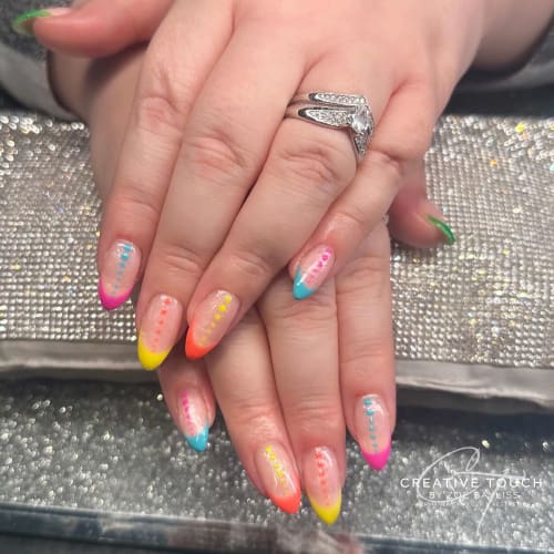 A close-up of hands with oval nails featuring a bright colour design with pink, yellow, orange, and blue tips and matching coloured dots running down the length of each nail.