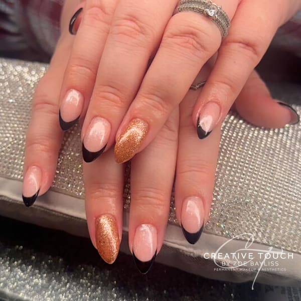 A close-up of hands with oval nails featuring a classic French tip design with black tips and a nude base, with the ring finger accented with glittery gold.
