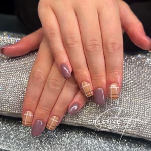 A close-up of hands with oval nails featuring a warm-toned tartan pattern in shades of brown, beige, and taupe.