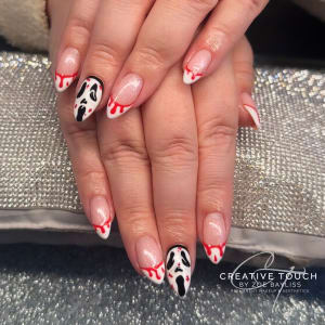 A close-up of hands with almond-shaped nails featuring a French tip design aaccented with red blood drip details and a black and white Scream mask on the middle finger.