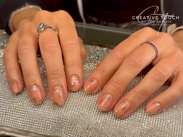 A close-up of hands with oval nails featuring a nude base coat accented with gold glitter.
