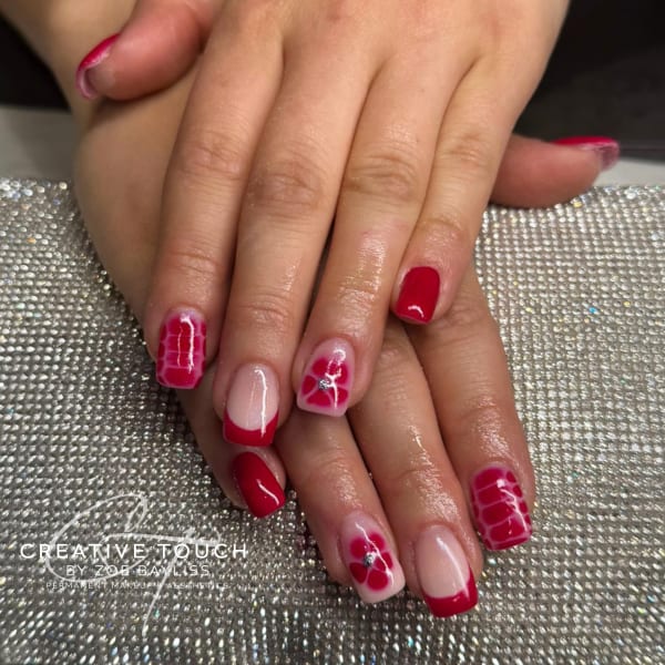 Close-up of hands with short, square-oval nails featuring a vibrant red and nude base. Designs include solid red nails, a red French tip, a red textured tortoiseshell pattern, and a delicate flower with a rhinestone centre on a nude base.