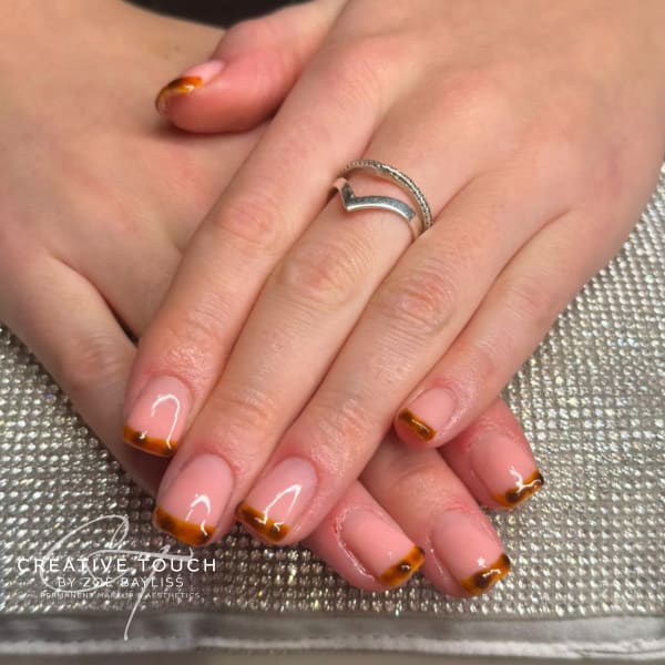 Close-up of hands with short, square-oval nails featuring a French tip design in warm brown with darker, subtle spots, giving a hint of tortoiseshell or abstract leopard print on a nude base.