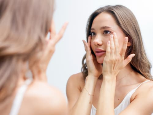 A warmly lit close-up of a woman standing in front of a large mirror, gently applying a skincare product (such as a tallow balm) to her face, conveying a sense of mindful self-care and attention to skin health.