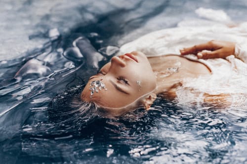 A beautiful young woman in a flowing dress lies partly submerged in the water. Fashion photography.