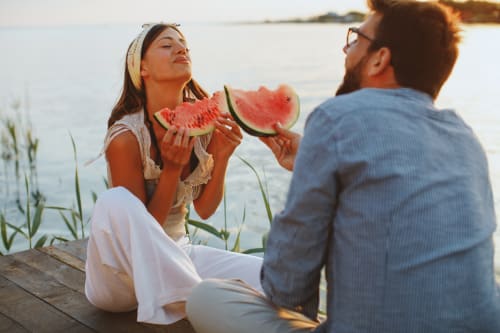 A young couple eating watermelon whilst sat by a river.