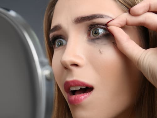 A woman looking distraught as she looks into a small mirror whilst pulling out loose eyelash extensions.