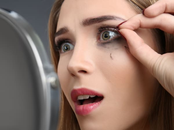 A woman looking distraught as she looks into a small mirror whilst pulling out loose eyelash extensions.