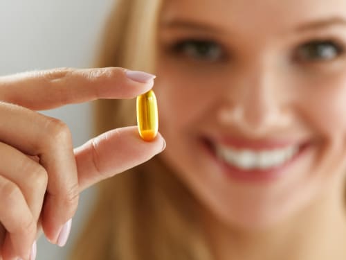 An attractive young woman with great skin holds a vitamin pill up to the camera whilst smiling in the background. Short field of view with focus on the vitamin pill.s