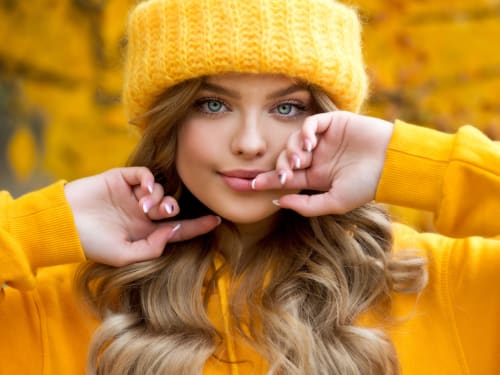 A beautiful young woman with perfect skin, dressed in amber coloured clothes and woolly hat. In the background are trees with autumn leaf colours.