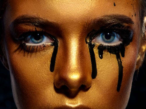 Pollution and health concept: a close-up portrait focused on the eyes of a woman in gold body paint. Black ink flowing from the corners of her eyes.