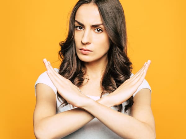An attractive woman standing in front of a plain orange background. She is looking stern with her arms crossed in front of her, signifying 