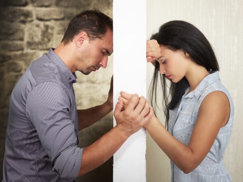A man and woman stand divided by an intimacy barrier between them. Each looks down in sadness as they hold hands to one side of the wall.