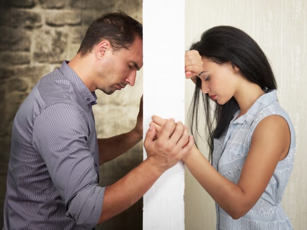 A man and woman stand divided by an intimacy barrier between them. Each looks down in sadness as they hold hands to one side of the wall.