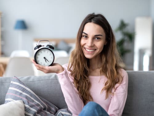 A smiling woman sitting on her sofa holding a clock.