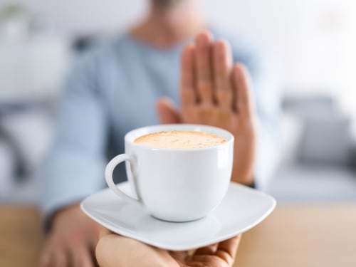 A person's outstretched hand pushes away a white cup of coffee offered on a saucer, indicating refusal or avoidance.