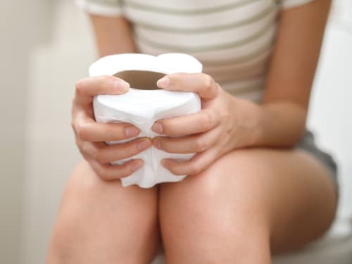 A woman with constipation sitting, clutching a toilet roll in the bathroom.