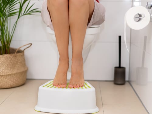 A person's bare feet are positioned on a small white step stool in front of a toilet, elevating their knees to promote a more natural squatting posture.