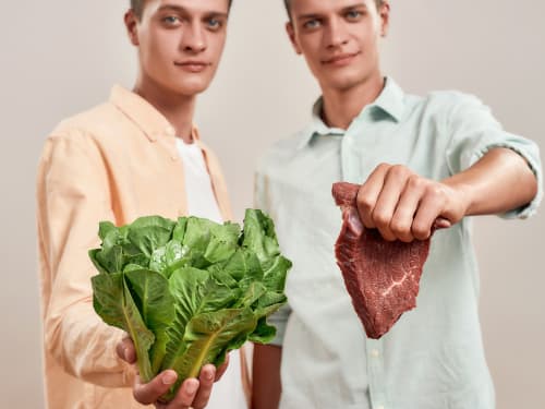 Twins, one holding a large bunch of leafy green vegetables and the other holding a raw cut of steak, against a plain background.