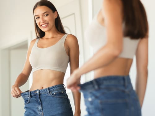 A smiling young woman looking at her reflection in a mirror, celebrating her weight loss journey