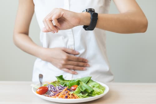 A person wearing a white shirt holds their hand to their stomach while checking a smartwatch, with a small plate of salad in the foreground, symbolising feelings of hunger and strict meal timing on a diet.