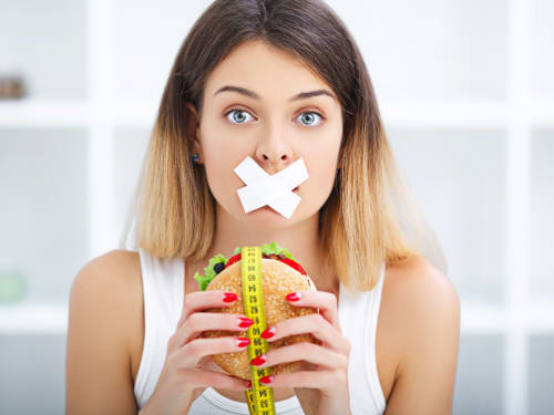 The conflict between diet restriction and intense food cravings: a woman with her mouth covered by tape, holding a burger wrapped in a yellow tape measure.