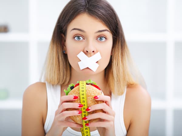 The conflict between diet restriction and intense food cravings: a woman with her mouth covered by tape, holding a burger wrapped in a yellow tape measure.