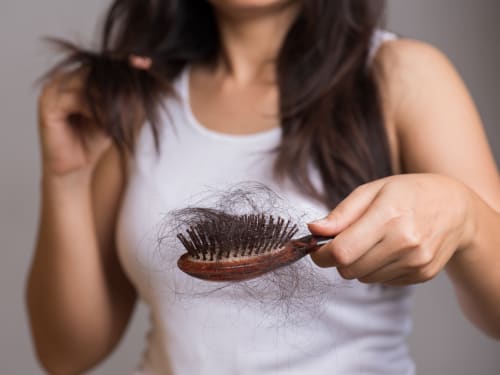 A woman holds out her hair brush to the camera, with bristles full of hair clumps. In the background she holds strands of hair.