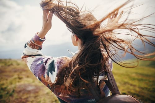 Lifestyle image showing a young woman with long, thick, windblown hair standing on top of a hill in the countryside