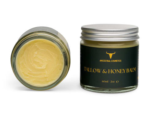 Ancestral Cosmetics Tallow & Honey Balm, 60ml/2oz jar. The product is shown with one jar standing upright, featuring a dark green label with a gold logo and text, and a silver lid. A second, identical jar is lying on its side, showing the creamy, pale yellow balm contents with visible texture inside the glass jar.