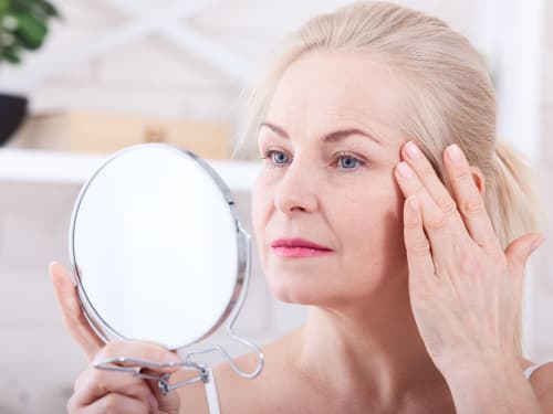 An older woman with blonde hair examines her facial skin closely using a handheld mirror,  touching her temple with her fingertips in a softly lit bathroom setting.