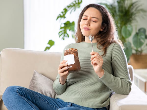 A woman relaxing on a sofa in the afternoon, eyes gently closed with a satisfied expression as she enjoys a spoonful of chocolate spread straight from the jar &mdash; capturing that familiar 4pm carb craving the article explains as a normal metabolic response, 