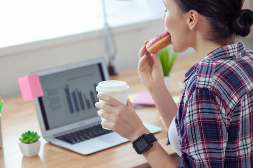 A woman at her desk in the mid-afternoon, taking a bite of a donut while holding a takeaway coffee, with a laptop showing work charts in the background &mdash; illustrating that familiar 4pm energy dip and sudden carb craving many people experience after lunch.