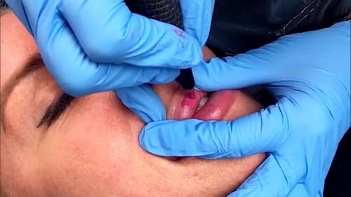Close-up shot of a red lip tattoo procedure in progress. A small needle deposits pigment into the client's lips.