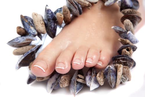 A wet woman's foot with French Pedicure nails rests on the ground with an anklet of muscle shells and driftwood around it.